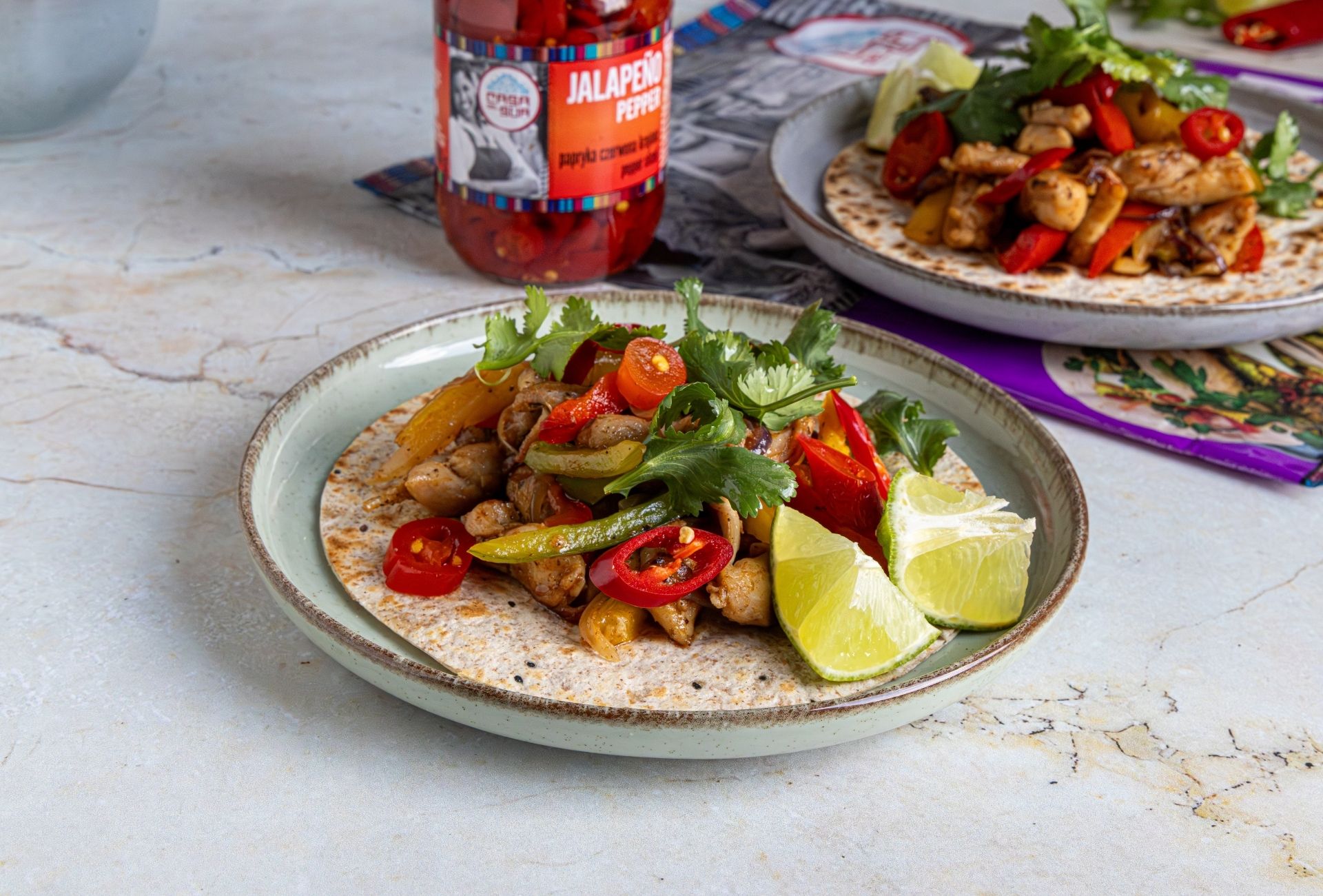 Fajita z kurczakiem, kolorową papryką, kolendrą i limonką podana na placku tortilla na talerzu, obok słoika papryczek jalapeño marki Casa del Sur.