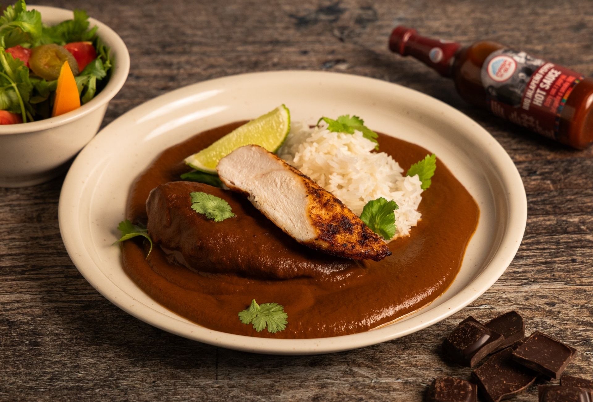 Pierś z kurczaka w sosie mole poblano z ryżem i limonką na talerzu, obok kawałków czekolady, sałatki oraz sosu BBQ Jalapeño Pepper marki Casa del Sur.