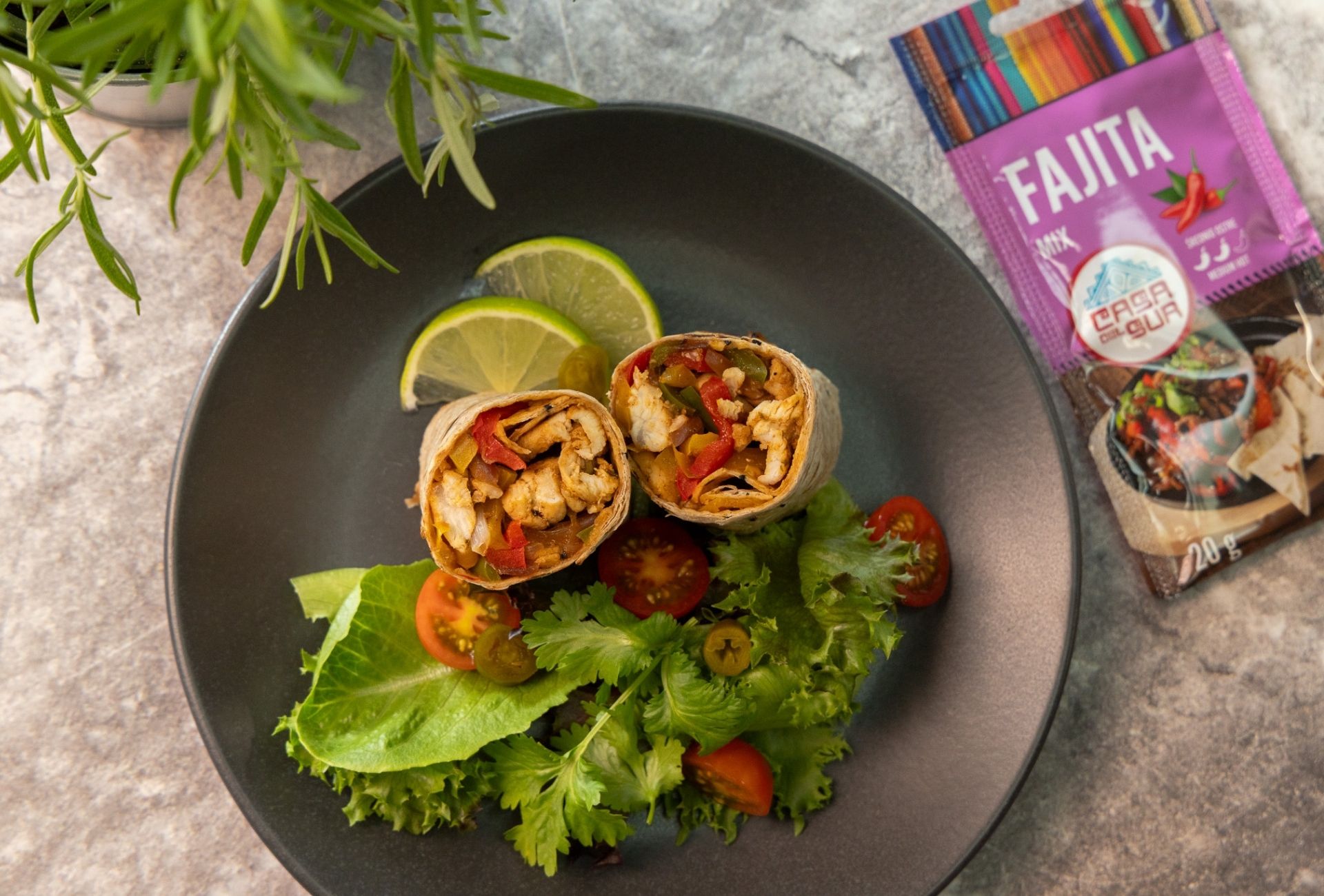 Przekrojone wrapy z kurczakiem fajitas, sałatą i limonką na czarnym talerzu, obok saszetki przyprawy Fajita Mix marki Casa del Sur.