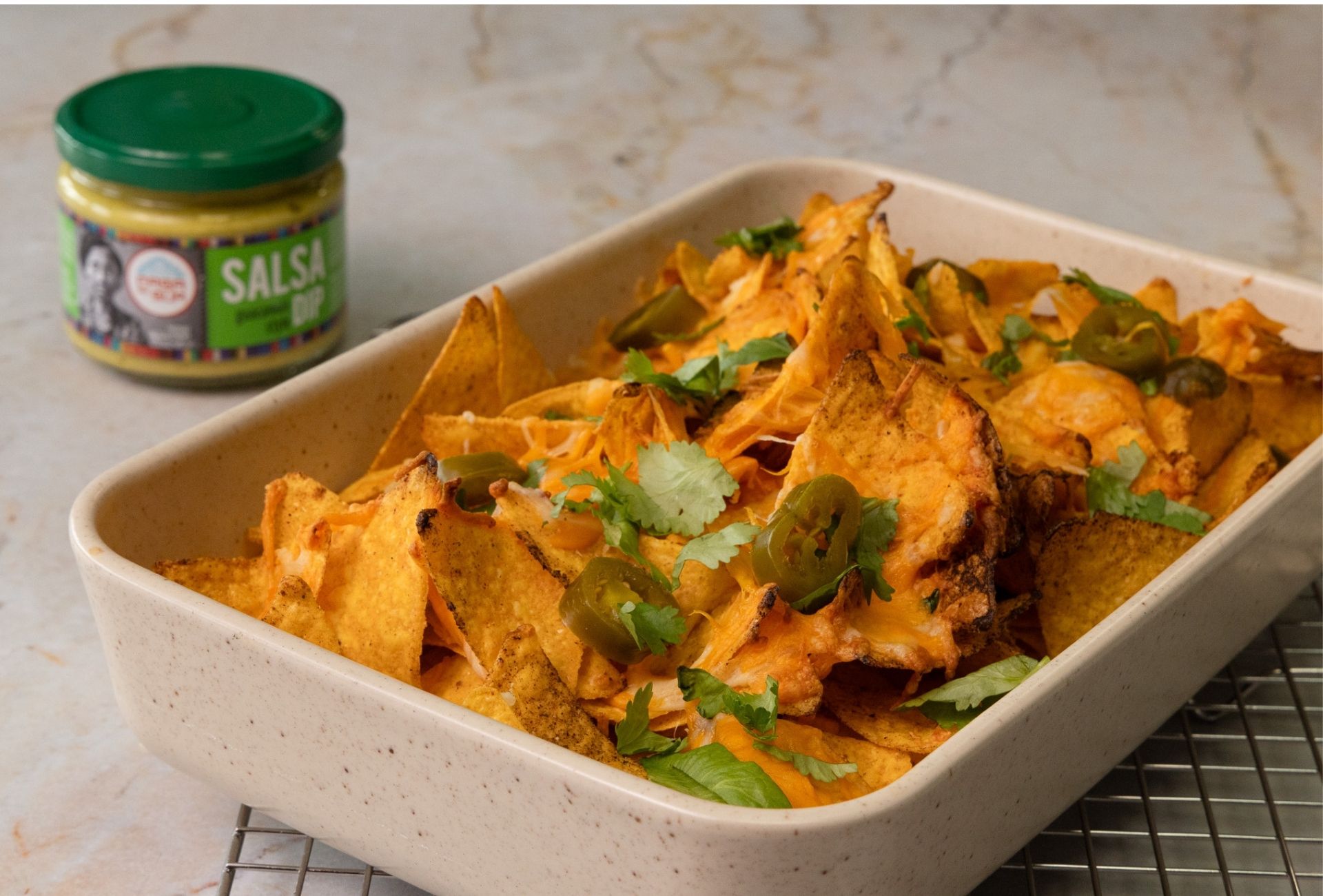 Chrupiące nachos zapiekane z serem, papryczkami jalapeño i kolendrą w prostokątnym naczyniu, podane obok słoiczka Salsa Dip z zieloną zakrętką marki Casa del Sur.