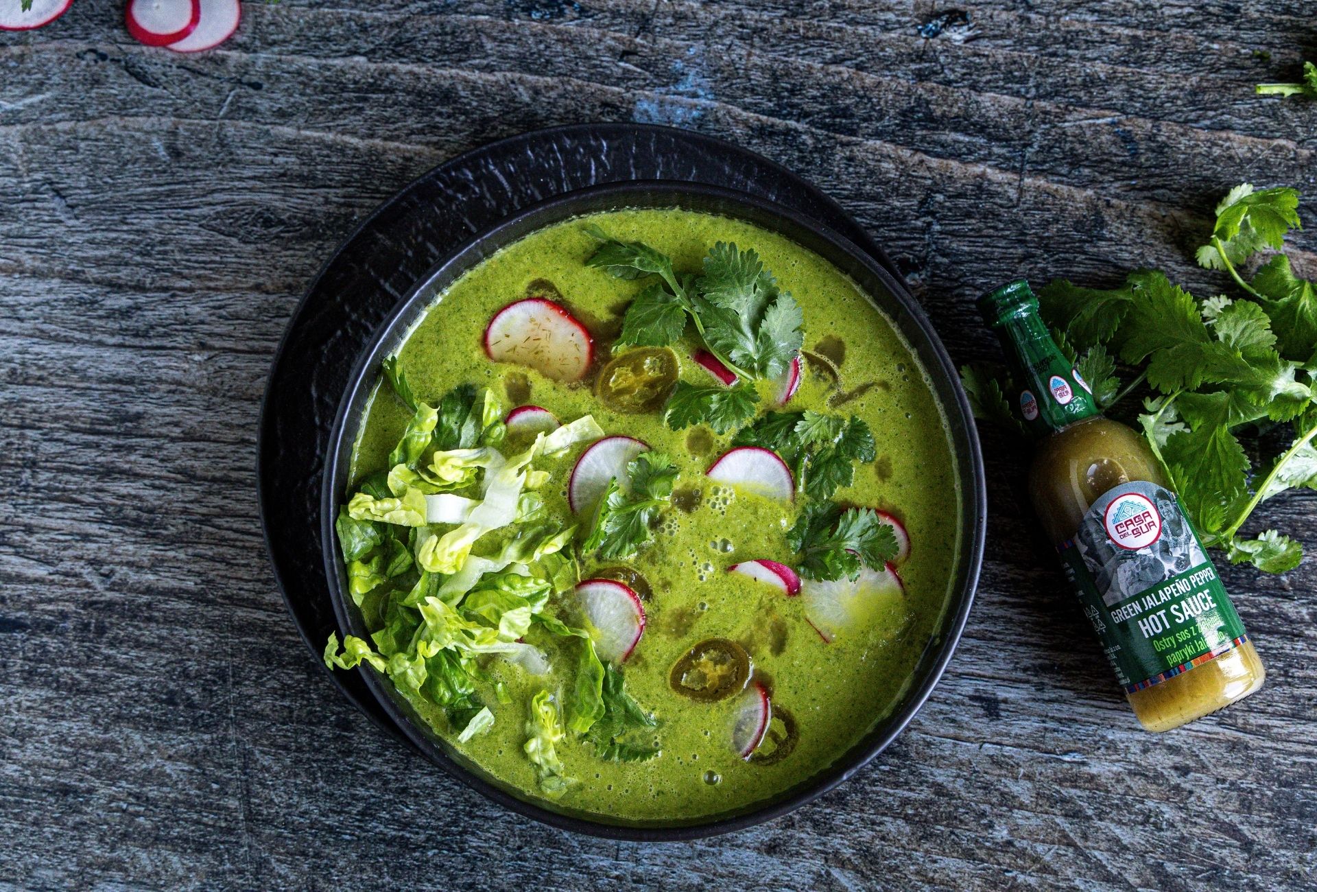 Zielona zupa Pozole verde z rzodkiewką, sałatą i kolendrą w czarnej misce, podana obok butelki sosu Green Jalapeño Pepper marki Casa del Sur.