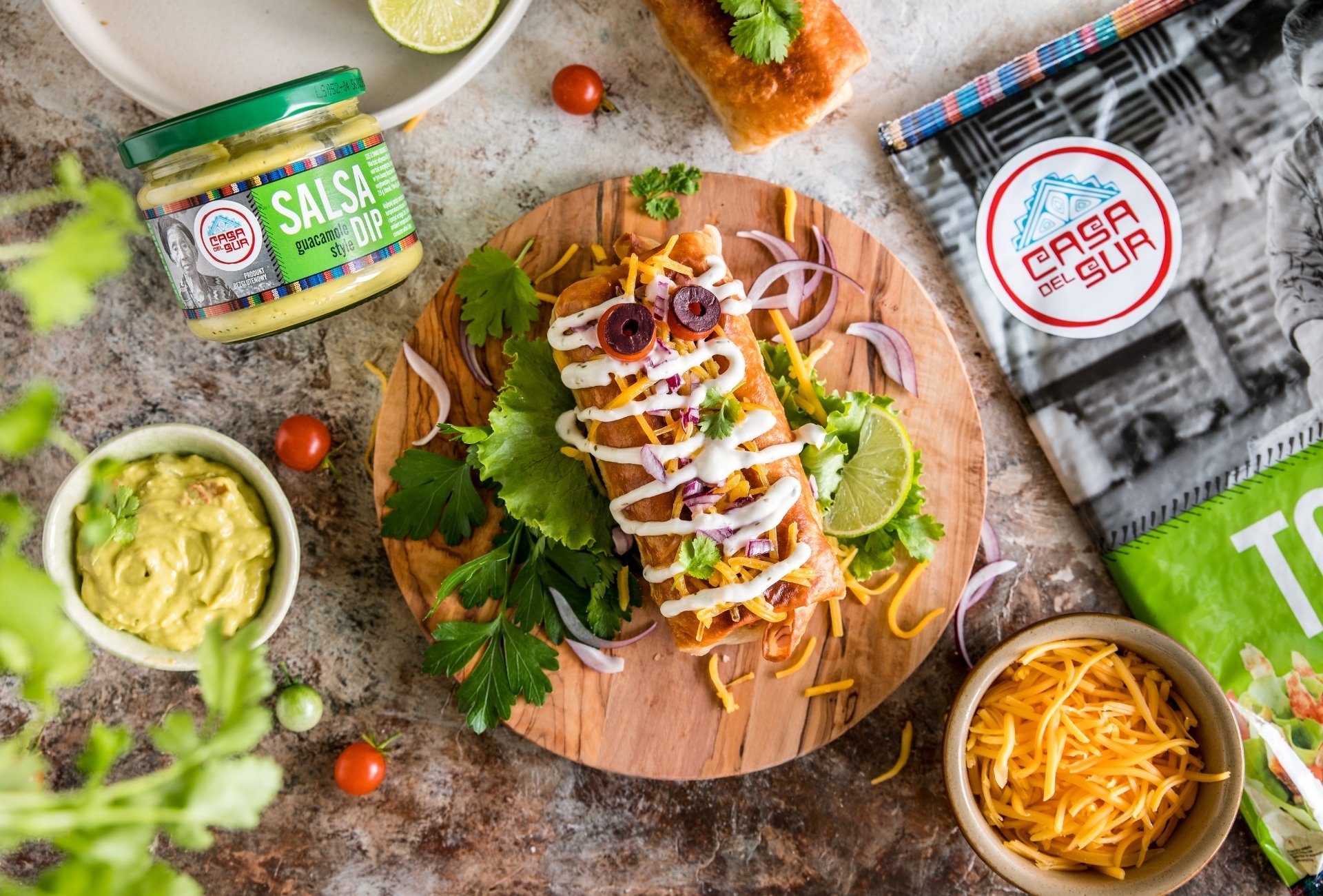 Chimichanga w kształcie mumii z oczami z oliwek i pomidorka, posypana serem cheddar, na tle produktów Casa del Sur