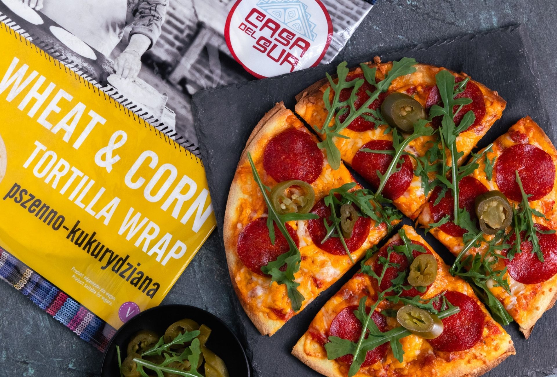 Zdjęcie przedstawia pizzę z salami i rukolą na tortilli, pociętą na kawałki i ułożoną na czarnej podkładce, obok żółtego opakowania wrapów „Casa del Sur”.
